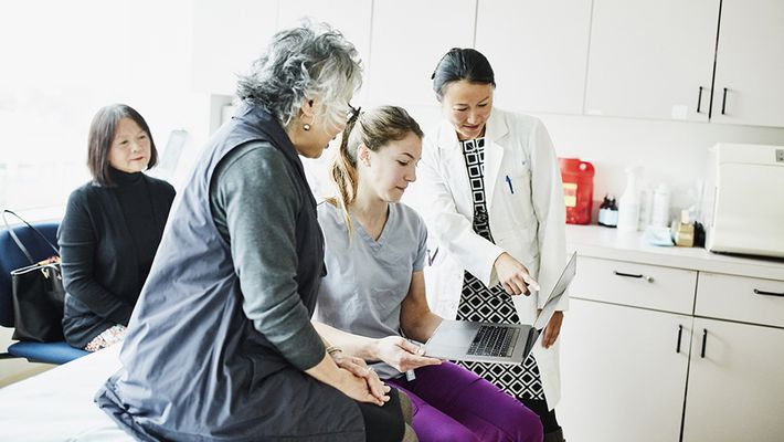 A doctor and nurse show a senior patient information on a laptop during a medical exam while another woman observes.